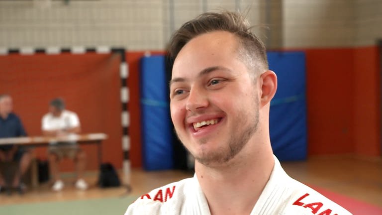 Ein junger Mann steht in einer Sporthalle und lächelt. (Foto: SWR)