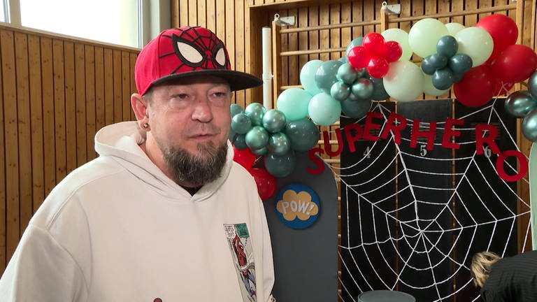 Ein Mann mit Spiderman-Kappe steht vor einer Sprossenwand mit Ballons und Superheldendekoration. (Foto: SWR)