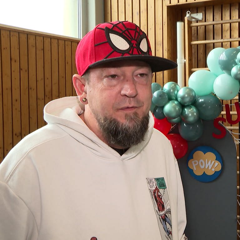 Ein Mann mit Spiderman-Kappe steht vor einer Sprossenwand mit Ballons und Superheldendekoration.