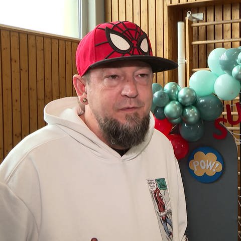 Ein Mann mit Spiderman-Kappe steht vor einer Sprossenwand mit Ballons und Superheldendekoration. (Foto: SWR)