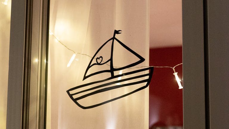 Ein Balkonfenster, beleuchtet mit einer Lichterkette und in der Mittel hängen die Konturen eines kleinen Segelbootes. Auf dem Segel ist ein Herz abgebildet. (Foto: SWR)