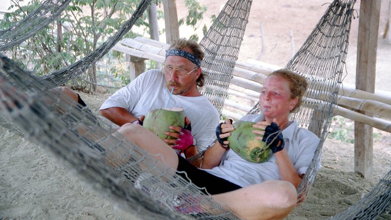 Zwei junge Menschen in einer Hängematte. Sie trinken aus einer Kokosnuss. 
