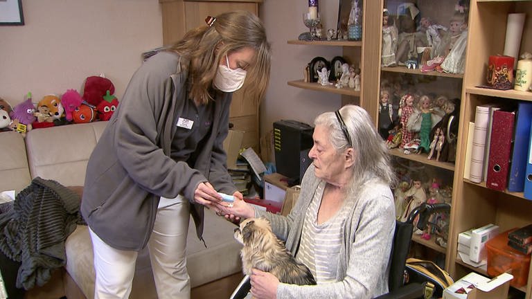 Eine junge Mitarbeiterin des Pflegedienstes versorgt eine ältere Dame mit Hund auf dem Schoß. 