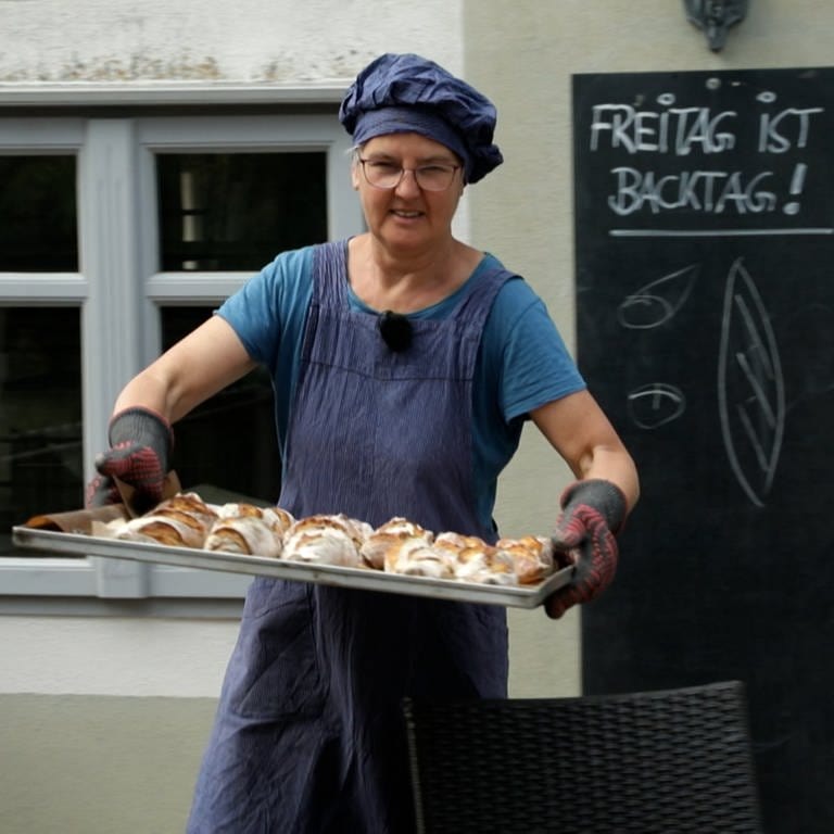 Frau mit blauer Mütze und Schürze trägt Blech mit Brot.