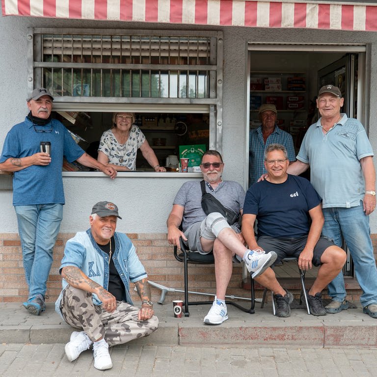 Eine Gruppe von Männern vor einem Kiosk, einige stehen, andere sitzen.