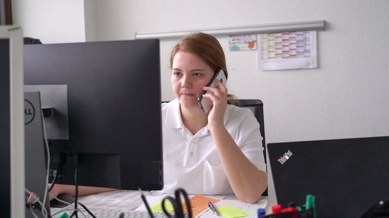 Junge Frau mit roten Haaren und weißem Polo-Shirt sitzt an einem Computer und telefoniert mit einem Handy.  (Foto: SWR)