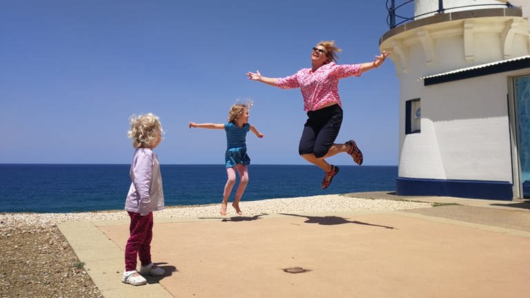 Ältere Dame springt mit Kindern in die Luft. Im Hintergrund das Meer und ein Leuchtturm.