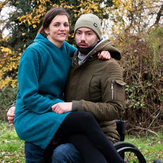 Eine lächelnde Frau sitzt auf dem Schoß ihres Mannes, der im Rollstuhl sitzt (Foto: SWR)