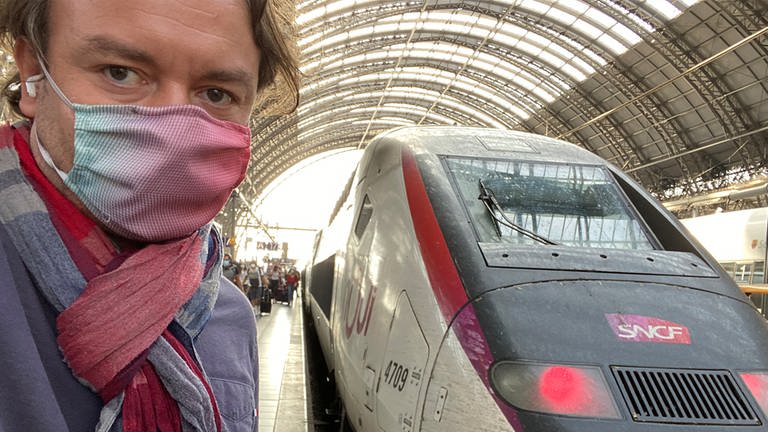 Mann mit Mundschutz steht vor einem weißen TGV im Bahnhof. 