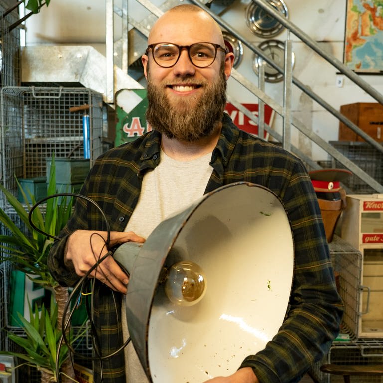 Chris, junger Mann mit Vollbart, in seiner Werkstatt. In der Hand eine große Lampe (Foto: SWR)