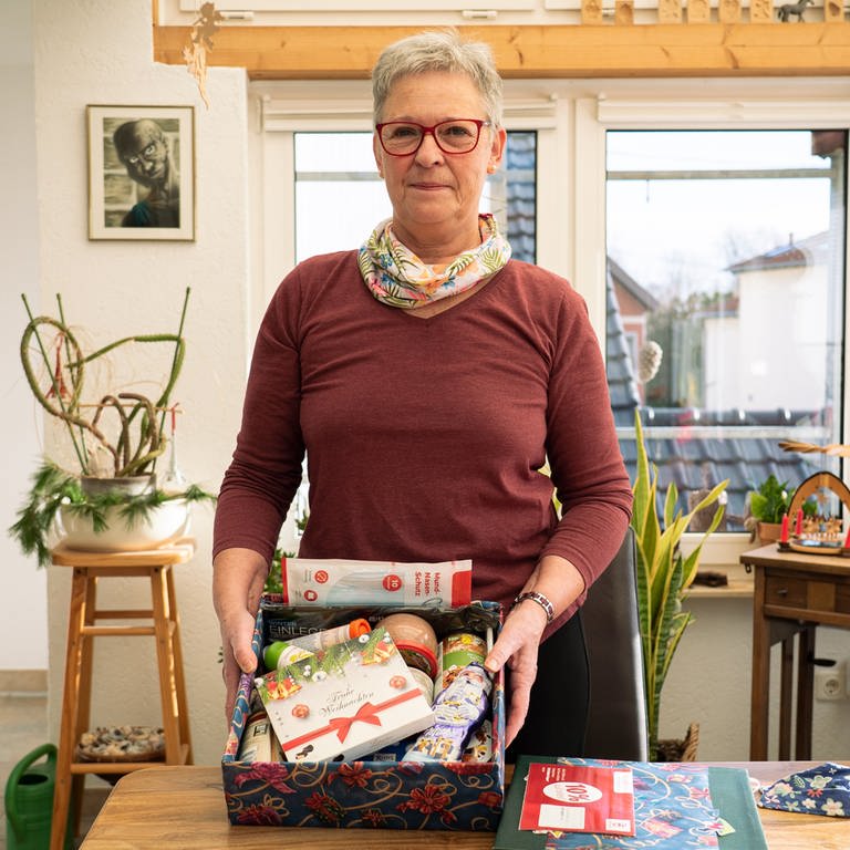 Ursula hat ein weihnachtliches Päckchen in der Hand. In dem Päckchen sind zum Beispiel Schokolade, Kaffee, warme Einlegesohlen und Wurstdosen.