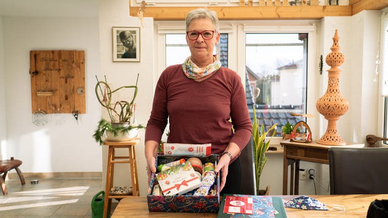 Ursula hat ein weihnachtliches Päckchen in der Hand. In dem Päckchen sind zum Beispiel Schokolade, Kaffee, warme Einlegesohlen und Wurstdosen. (Foto: SWR)
