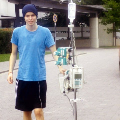 Maximilian steht neben einem Infusionsspender vor einer Klinik (Foto: SWR)