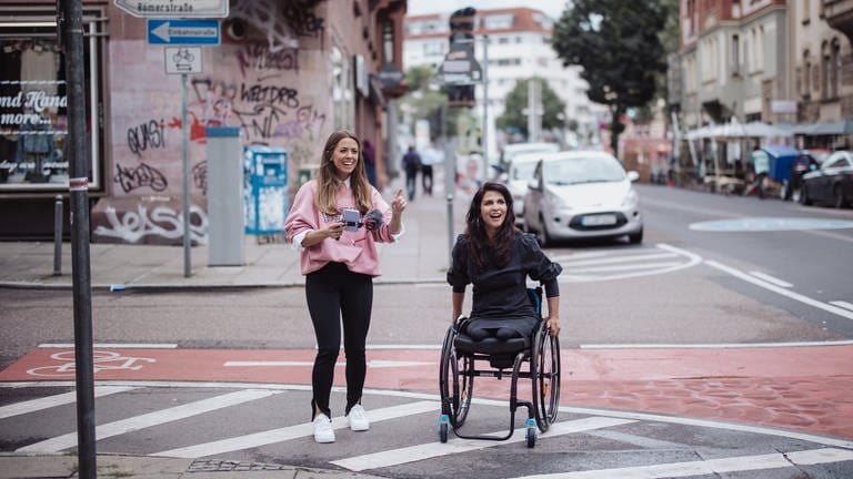 Zwei Frauen überqueren eine Straße. Eine sitzt im Rollstuhl, die andere läuft.