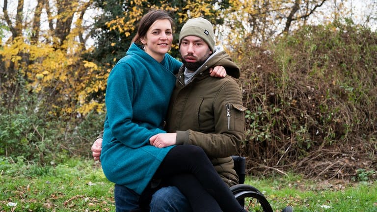 Eine lächelnde Frau sitzt auf dem Schoß ihres Mannes, der im Rollstuhl sitzt
