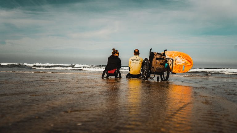 Mit dem Rücken zur Kamera: Der querschnittgelähmte Johannes Laing sitzt mit einer gleichaltrigen Frau am Strand und sie beobachten die Wellen.  (Foto: SWR)