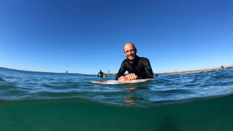 Der querschnittgelähmte Johannes Laing ist im Wasser, stützt sich auf sein Surfbrett und grinst in die Kamera. (Foto: SWR)