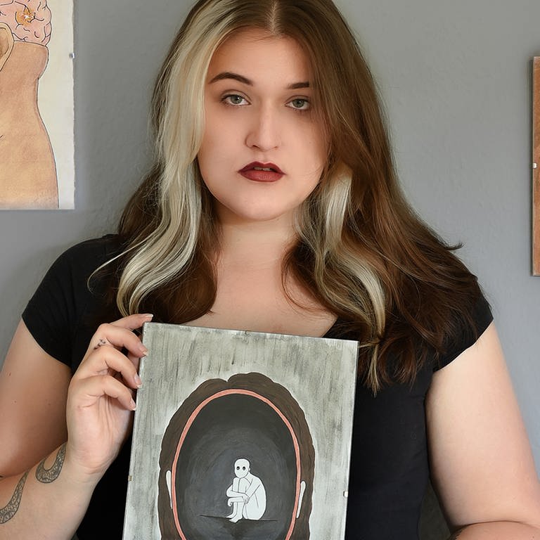 Junge Frau mit einem von ihr gemalten Bild in der Hand, vor einer Wand mit weiteren von ihr gemalten Bildern (Foto: SWR)