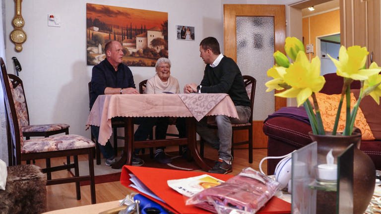 Mann, Anfang 50, bespricht sich mit Seniorin und einem ihrer Bekannten in ihrem Wohnzimmer (Foto: SWR)