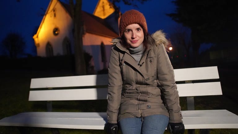 Junge Frau in Winterkleidung auf weißer Bank vor Kirche bei Nacht mit Blick in die Kamera