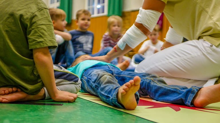 Ein Kind liegt auf einer Matte in der Turnhalle an der Goetheschule in Mainz. Mine Musahl zeigt hier den Vier- und Fünfjährigen, wie sie überprüfen, ob jemand bewusstlos ist. (Foto: SWR)
