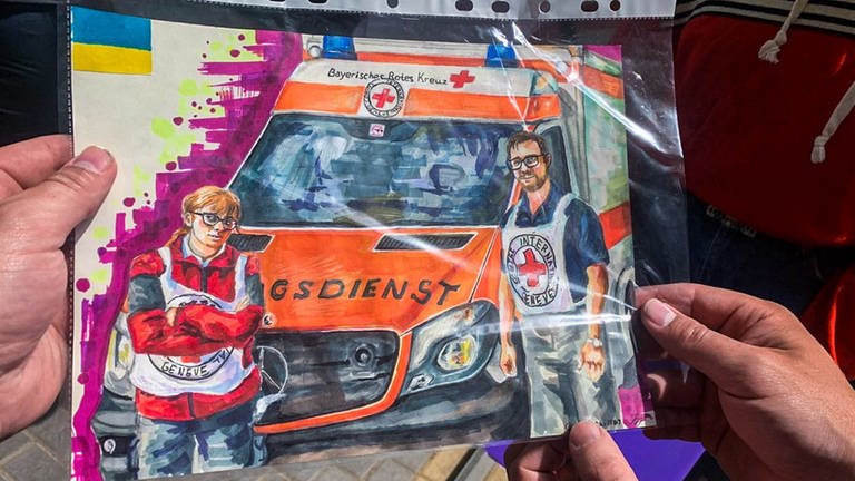 Ein gemaltes Bild zeigt Rouven und eine Kollegin vom Roten Kreuz vor einem Rettungswagen. Die Sanitäter waren vier Wochen in der Ukraine im Einsatz.