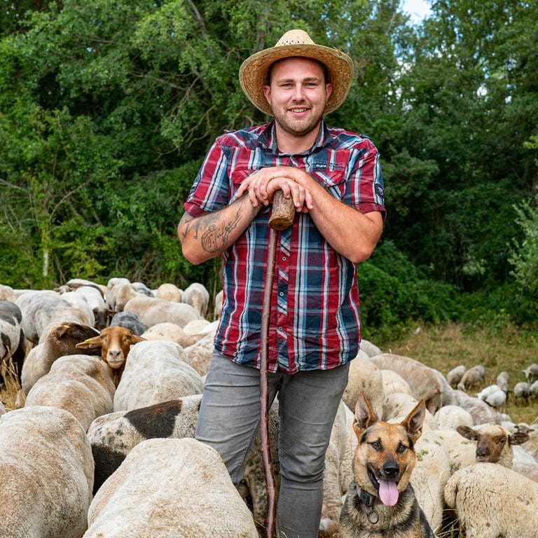 Junger Mann mit kariertem Hemd und Strohhut lehnt vor einem Wald auf einem Stock. Um ihm herum sind Schafe.  (Foto: SWR)
