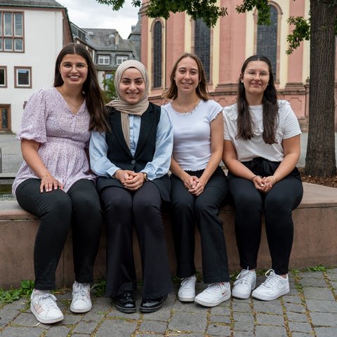 Vier junge Finanzbeamtinnen sitzen nebeneinander auf dem Rand eines Brunnens auf dem Wormser Marktplatz und lächeln. (Foto: SWR)