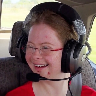 Junge Frau sitzt im Flugzeug, trägt Kopfhörer und lächelt.  (Foto: SWR)