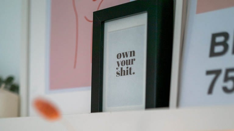 Auf einem eingerahmten Bild steht „own your shit” (Foto: SWR)