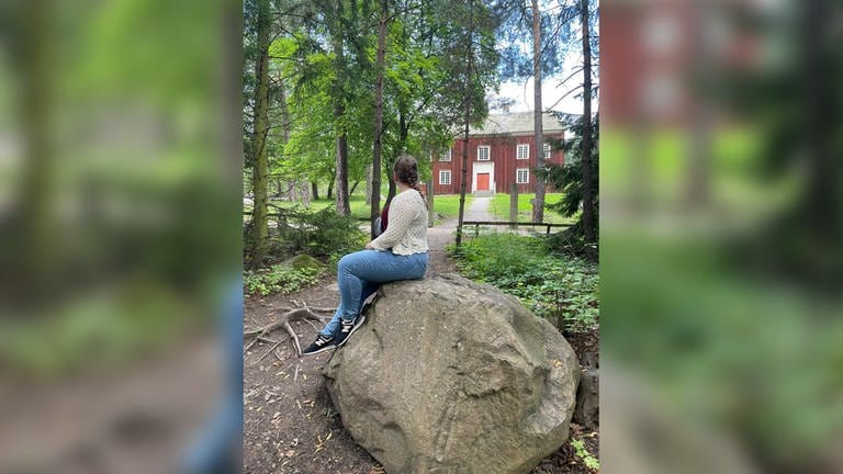 Eine junge Frau sitzt auf einem Stein. Im Hintergrund ist ein rotes Haus zu sehen. (Foto: SWR)