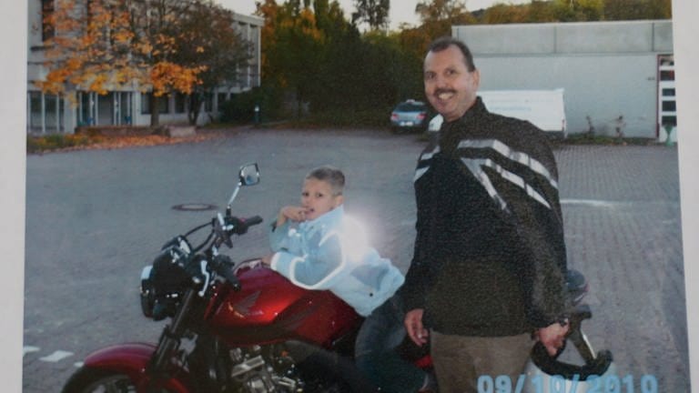 Ein Kind sitzt auf einem Motorrad daneben ein Mann mittleren Alters mit Motorradhelm. Der Mann lächelt in die Kamera.
