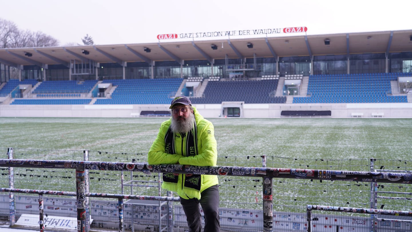 David trägt Football-Fanartikel der Stuttgart Surge. Er ist im GAZi-Stadion (Stuttgart Surge Stadion) und steht im B-Block. Er lehnt sich über ein Geländer und schaut in die Kamera. Im Hintergrund ist das Stadion mit der Haupttribüne zu sehen. (Foto: SWR)
