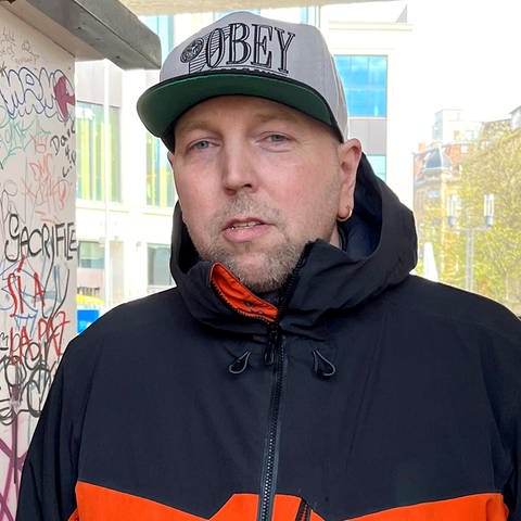 Simon steht neben dem Stand vom „Paule Club” in Stuttgart unter der Paulinenbrücke. Er ist Sprecher und Mitgründer des „Paule Clubs”. Er trägt eine graue Cap und eine schwarz-orangefarbene Jacke. Er schaut in die Kamera. 