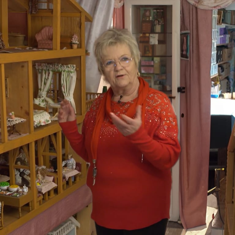Eine ältere Frau steht vor einem Puppenhaus und erzählt von ihrem besonderen Handwerk.