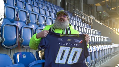 David trägt Football-Fanartikel der Stuttgart Surge. Er sitzt auf der Haupttribüne im GAZi-Stadion in Stuttgart und lächelt in die Kamera. Er zeigt stolz ein Trikot der Stuttgart Surge in die Kamera, auf dem die Football-Spieler unterschrieben haben. (Foto: SWR)