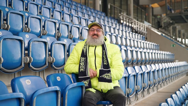 David trägt Football-Fanartikel der Stuttgart Surge. Er sitzt auf der Haupttribüne im GAZi-Stadion (Stuttgart Surge Stadion) in Stuttgart und lächelt in die Kamera. (Foto: SWR)