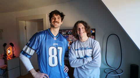 Content-Creator Zack Bachelor steht links im Bild und Zac Ryan rechts im Bild. Beide tragen Football-Fanartikel der Detroit Lions. Sie lachen in die Kamera, während im Hintergrund im Fernseher ein NFL-Football-Spiel läuft.