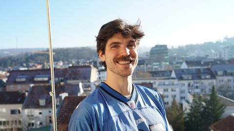 Content Creator und Footballliebhaber Zack Bachelor steckt auf einem Balkon und lächelt in die Kamera. Er trägt Football-Fanartikel der Detroit Lions. Im Hintergrund sind andere Wohngebäude Stuttgarts zu sehen.
