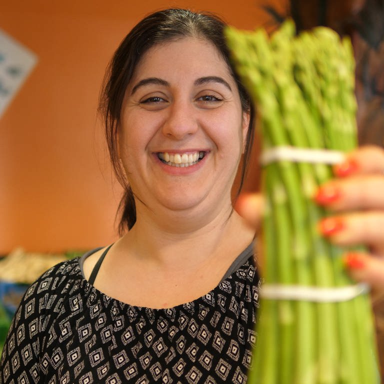 Noura hält einen bund grünen Spargel in die Kamera und lächelt dabei. (Foto: SWR)