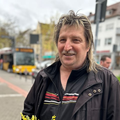 Michael steht in Bad Cannstatt mit Blumen und einer Topfpflanze in der Hand. Er lächelt in die Kamera. Im Hintergrund ist der Stuttgarter Verkehr zu sehen.