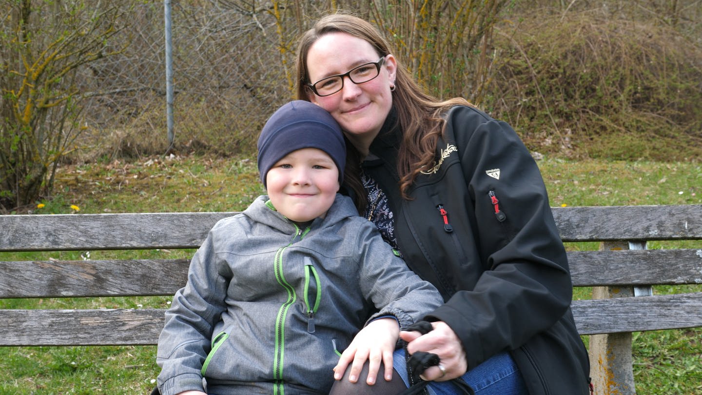 Eine junge Mutter sitzt auf einer Parkbank und lächelt zusammen mit ihrem Sohn in die Kamera. (Foto: SWR)