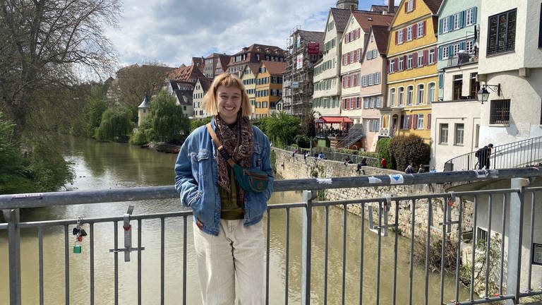 Laura steht auf der Eberhardsbrücke in Tübingen. Sie hat eine Jeansjacke an und ihre Hände in den Taschen. Hinter ihr sieht man Tübingen mit dem Hölderlinturm. 