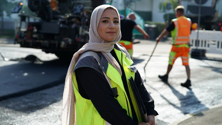 Bauingenieurin Hawra lächelt in die Kamera und steht auf einer Baustelle. Im Hintergrund sind Straßenbauarbeiter zu sehen.