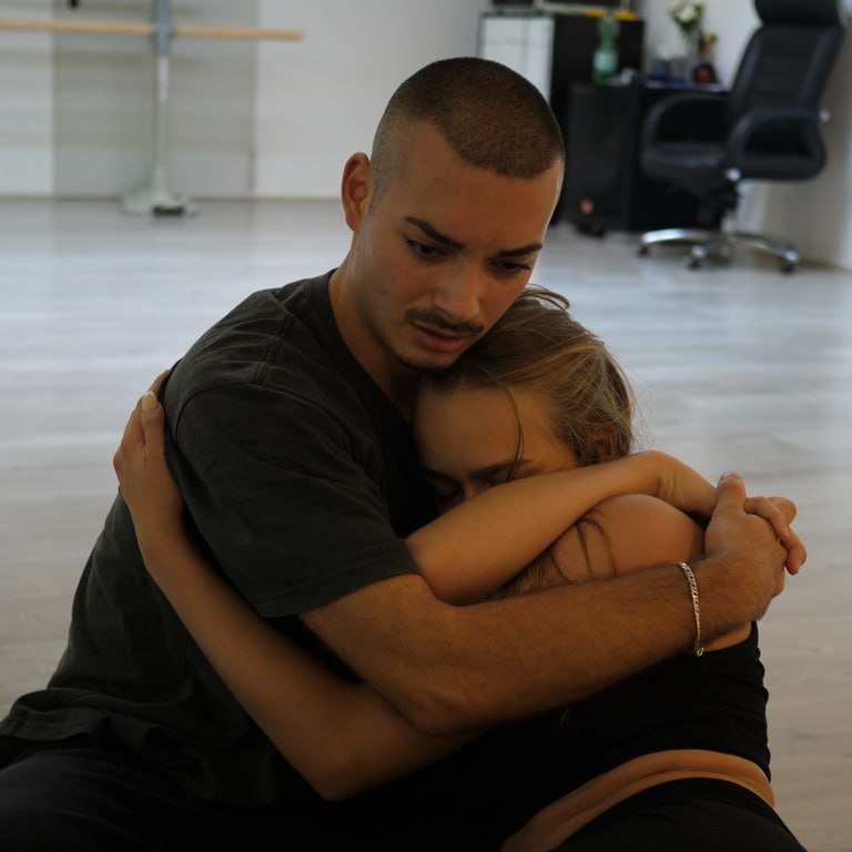 Tanzlehrer Deniz tanzt zusammen mit einer Tanzpartnerin. Die beiden sitzen auf dem Boden und halten sich in den Armen. Deniz’ Blick ist gefühlvoll und emotional.