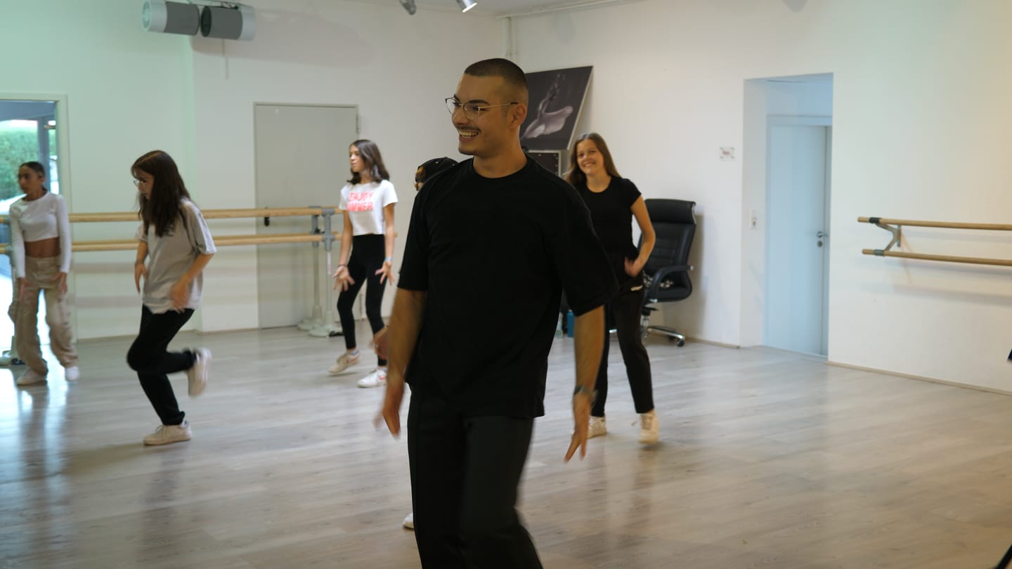 Tanzlehrer Deniz ist in einem Tanzstudio. Dort bringt er Kindern das Tanzen bei. Deniz macht eine Tanzübung vor und lacht dabei. Im Hintergrund sind Kinder zu sehen, die Deniz’ Tanzbewegung nachmachen. (Foto: SWR)