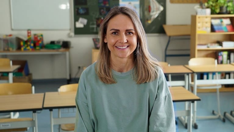 Eva-Maria ist Trainerin für sensomotorische Integration und fördert die Resilienz von Kindern an Schulen. Sie sitzt auf einem Tisch in einem Klassenzimmer. Im Hintergrund sind Schulutensilien und eine Tafel zu sehen. Eva-Maria trägt ein grünes Oberteil und lächelt in die Kamera.