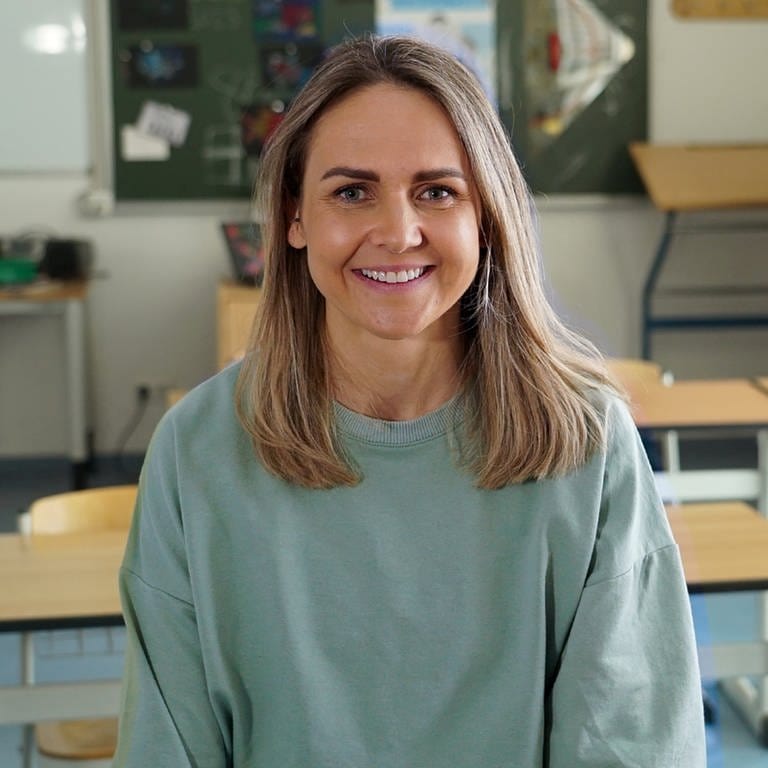 Eva-Maria ist Trainerin für sensomotorische Integration und fördert die Resilienz von Kindern an Schulen. Sie sitzt auf einem Tisch in einem Klassenzimmer. Im Hintergrund sind Schulutensilien und eine Tafel zu sehen. Eva-Maria trägt ein grünes Oberteil und lächelt in die Kamera.