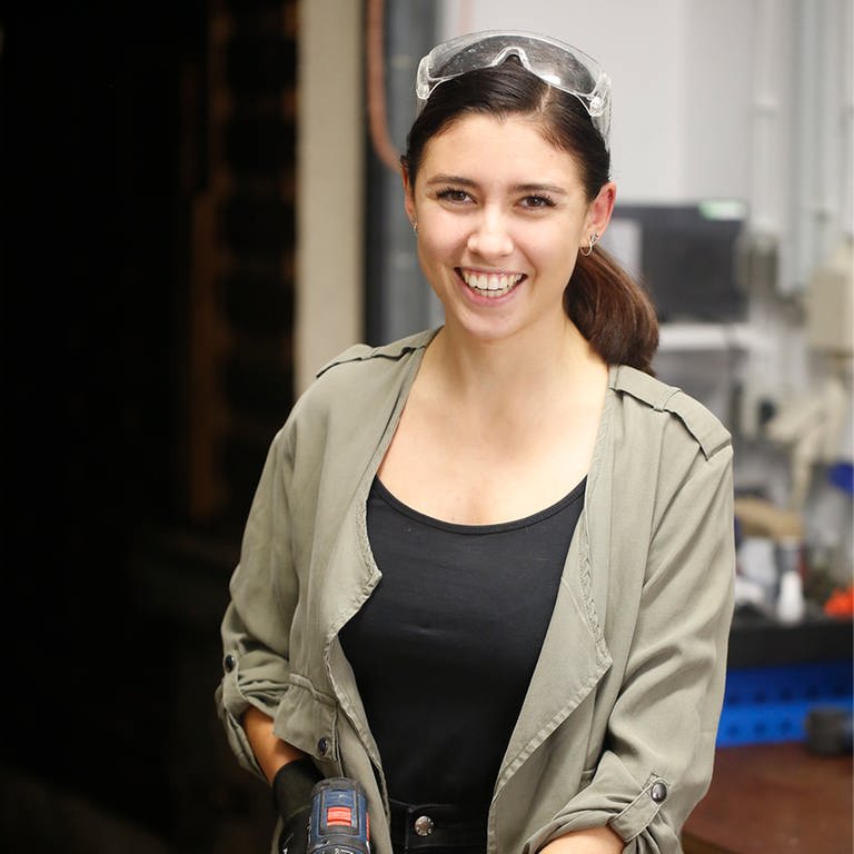 Junge Frau steht mit Schutzbrille und Bohrmaschine in einer Werkstatt und lächelt in die Kamera