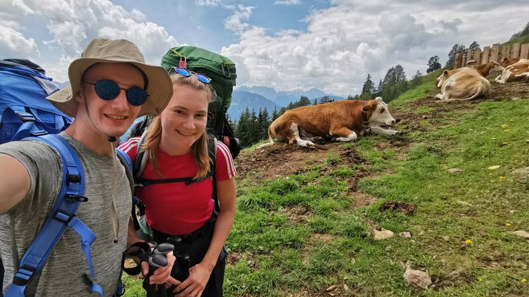 Zwanzigjähriger Frau und Mann stehen an einem Berg. Im Hintergrund liegen mehrere Kühe. 